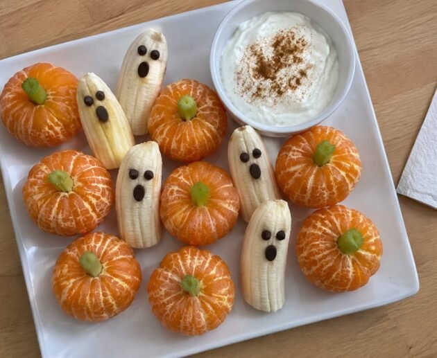 Plato casero con mandarinas peladas como calabacitas con tallo de apio, plátanos fantasma con ojos de chocolate y dip de yogur con canela