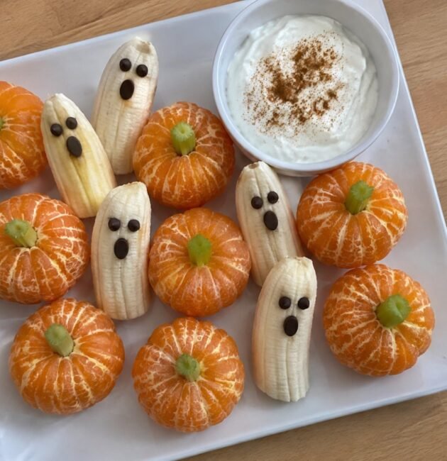 Plato casero con mandarinas peladas como calabacitas con tallo de apio, plátanos fantasma con ojos de chocolate y dip de yogur con canela
