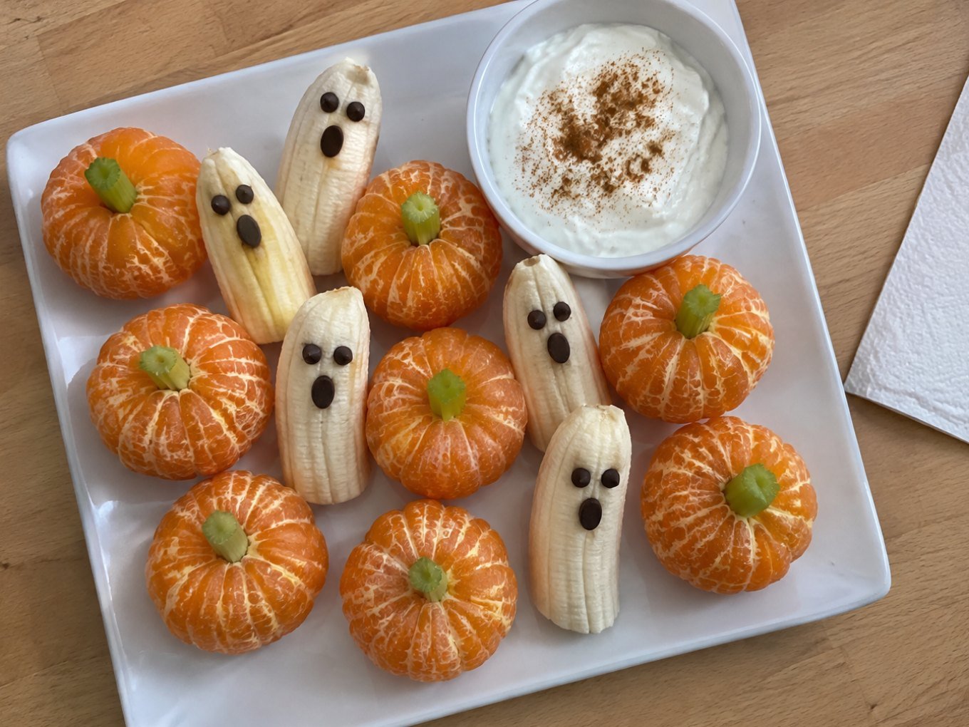 Plato casero con mandarinas peladas como calabacitas con tallo de apio, plátanos fantasma con ojos de chocolate y dip de yogur con canela