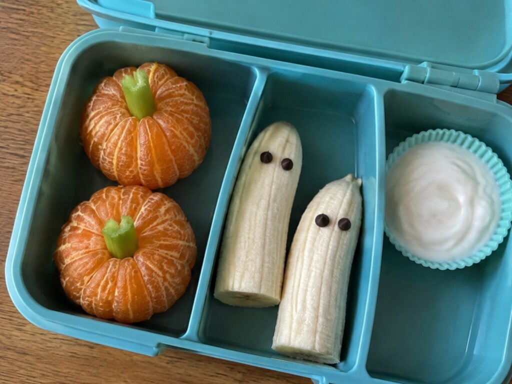 Lonchera escolar abierta con mandarinas calabaza, plátanos fantasma y vasito de dip de yogur, idea de lunch de Halloween saludable para niños