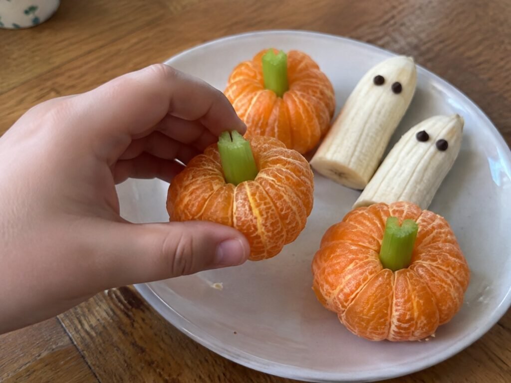 Mano de niño tomando una mandarina calabaza con tallo de apio de un plato con plátanos fantasma en una mesa de madera casera