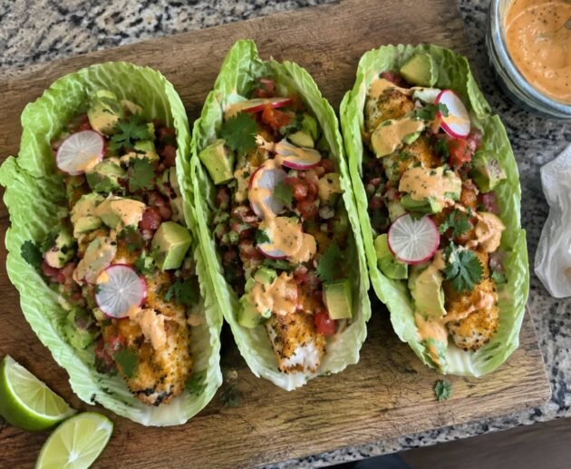 Tres tacos en hoja de col con pescado empanizado al horno, aguacate, pico de gallo y aderezo de chipotle sobre tabla de madera