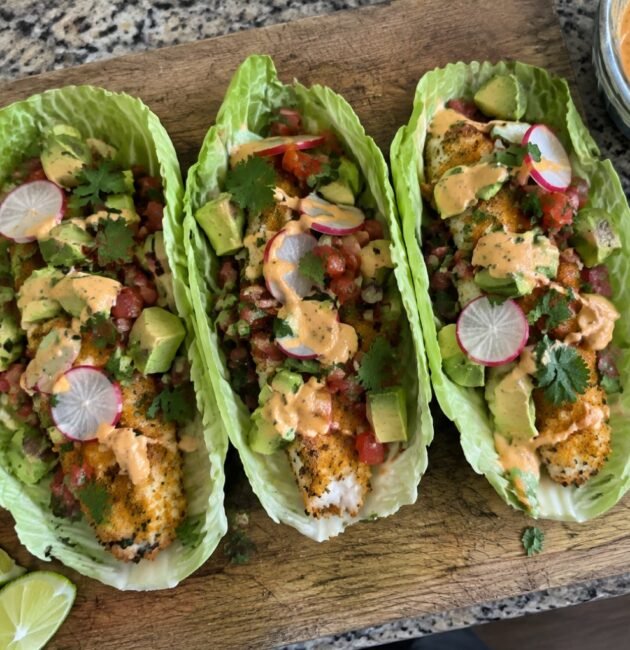 Tres tacos en hoja de col con pescado empanizado al horno, aguacate, pico de gallo y aderezo de chipotle sobre tabla de madera