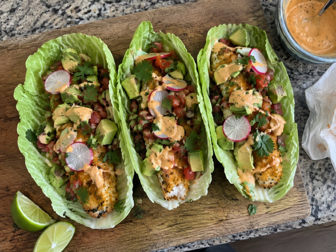Tres tacos en hoja de col con pescado empanizado al horno, aguacate, pico de gallo y aderezo de chipotle sobre tabla de madera