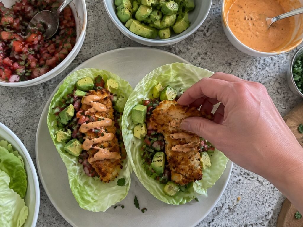 Manos armando tacos en hojas de col con tiras de pescado al horno, pico de gallo, aguacate y aderezo de chipotle en la barra