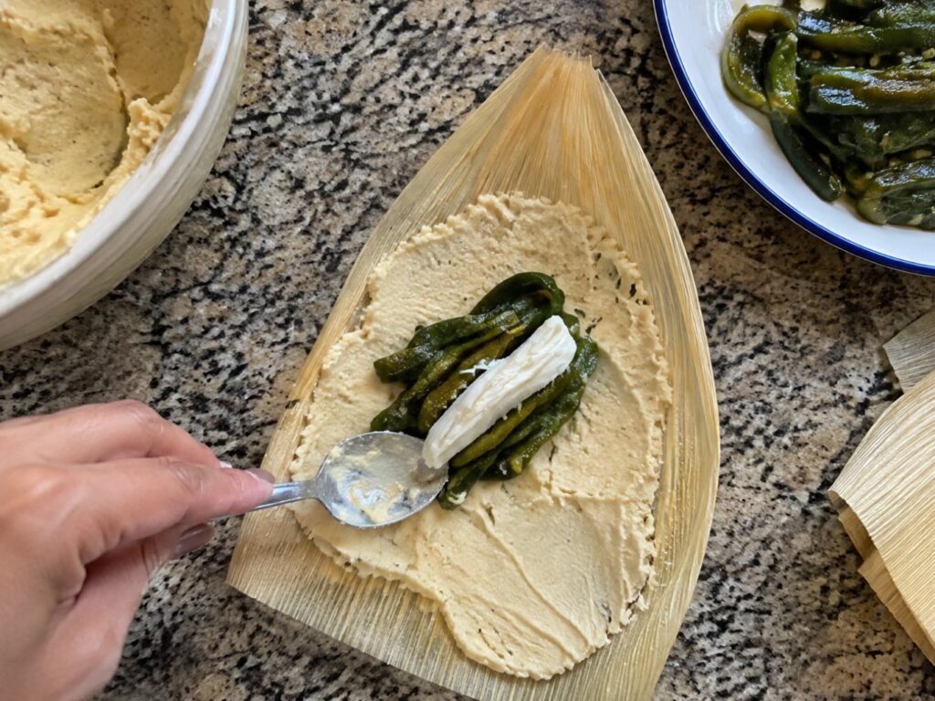 Mano untando masa sin manteca sobre hoja de maíz con rajas de poblano y queso, paso a paso para hacer tamales de rajas en casa