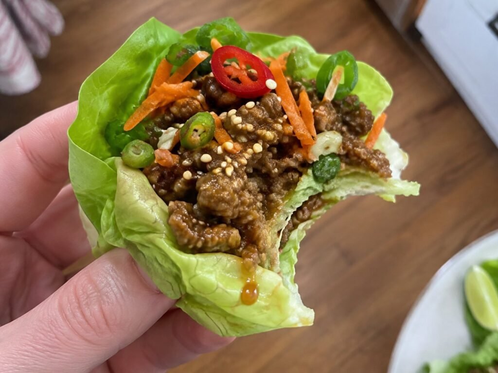 Mano sosteniendo un wrap de lechuga crujiente con carne molida jugosa en salsa asiática, zanahoria y ajonjolí, con una mordida