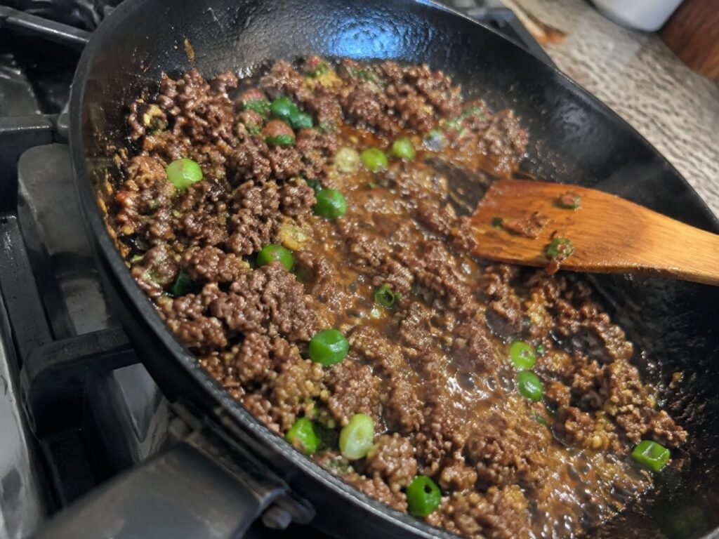 Carne molida dorándose en sartén negro con salsa de soya brillante, ajo, jengibre y cebolla cambray, paso fácil de la receta