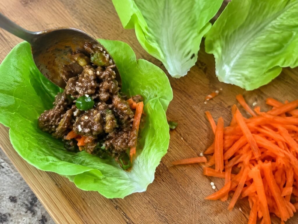 Cuchara colocando carne molida glaseada en una hoja de lechuga sobre tabla de madera junto a zanahoria rallada, armando wraps saludables